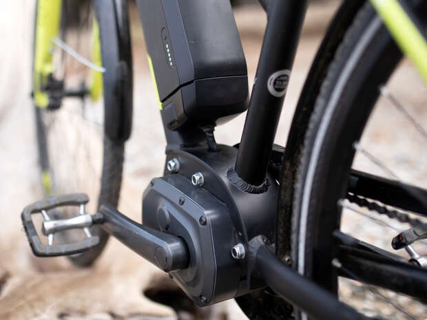 The electric bike's motor and battery mount near the pedals, with a few LED indicators lit. It's on a paved path, surrounded by greenery and sunlight.