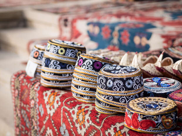 Stacks of intricately embroidered hats rest on a vibrant patterned carpet, surrounded by other colorful textiles in an outdoor market setting.