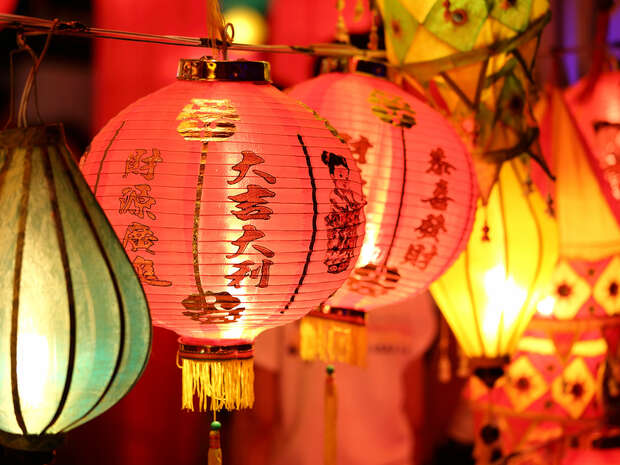 Colorful paper lanterns hang in a vibrant setting. The central lantern glows red with vertical black Chinese characters. Surrounding lanterns emit warm light, creating a festive atmosphere.