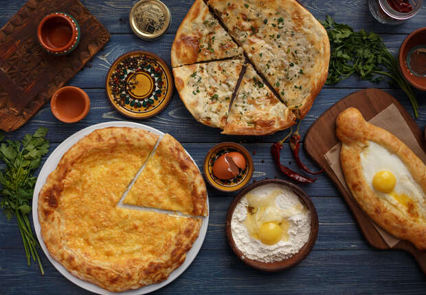 Pastries with cheese and eggs on a rustic table; nearby, there are small bowls of flour, egg yolks, and decorative plates, surrounded by fresh herbs and spices.
