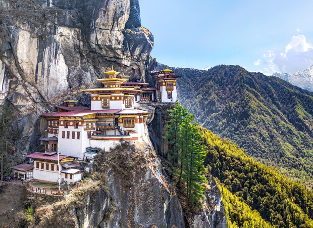 A multi-tiered, traditional monastery clings to a steep cliffside, overlooking a lush, forested valley with distant mountains under a clear sky.
