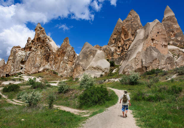 A person walks along a dirt path surrounded by unique rock formations with cave-like openings under a bright blue sky with scattered clouds. Sparse greenery lines the path.