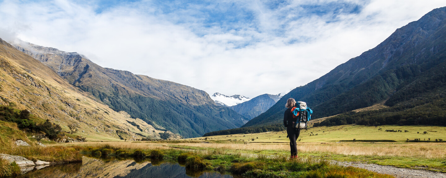 A backpacker stands on a grassy field, gazing at a mountainous landscape with a clear sky and a small pond reflecting the scenery beside them.