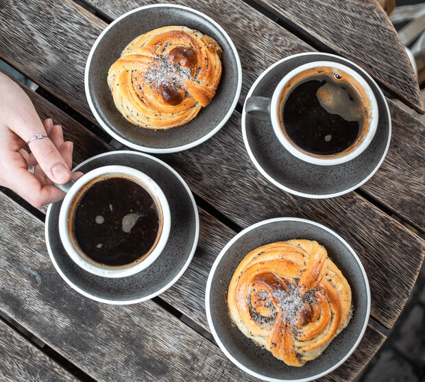 Cups of coffee sit on saucers beside sugar-dusted pastries, placed on a rustic wooden table. A hand reaches to pick up one coffee cup.