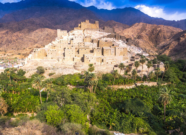 Ancient stone village sits perched on a rocky hillside, surrounded by lush greenery and palm trees, with mountainous terrain in the background under a partially cloudy sky.