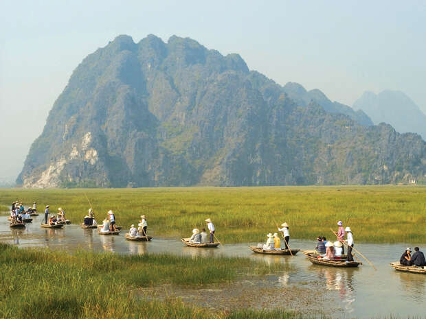 Wooden boats glide along a narrow waterway, with people rowing and riding, set against a backdrop of grassy wetlands and towering, misty mountains.