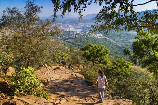 A person walks down a rocky path, surrounded by trees and foliage, overlooking a vast landscape with distant mountains and a town below under a clear blue sky.