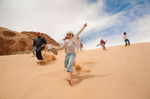 A group of barefoot people joyfully run down a sandy dune under a blue sky with scattered clouds, near rocky formations in a desert environment.