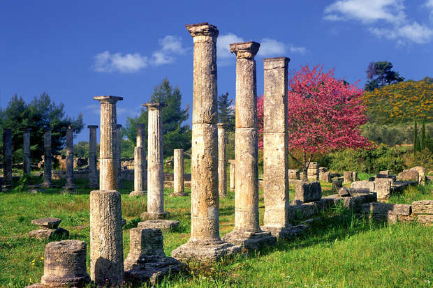 Ancient stone columns stand among ruins, surrounded by green grass and trees. A vibrant pink flowering tree adds color, with a backdrop of hills and a bright sky.