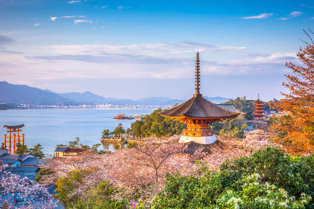 A pagoda stands among cherry blossoms overlooking a tranquil coastal scene with a floating torii gate and distant hills under a clear blue sky.