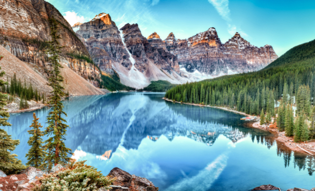 A clear, reflective lake mirrors rugged, rocky mountains under a blue sky, surrounded by dense evergreen forests, creating a serene and majestic natural landscape.