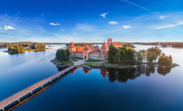 A castle with red roofs rests on an island, surrounded by a lake. A wooden bridge connects the island to the mainland. Trees and a blue sky complete the scene.