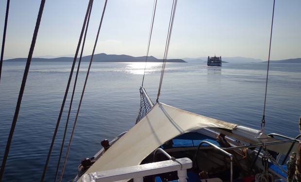 A boat with a sunshade at the bow is docked on calm water. In the distance, another vessel sails towards the horizon, with silhouetted islands under a clear sky.