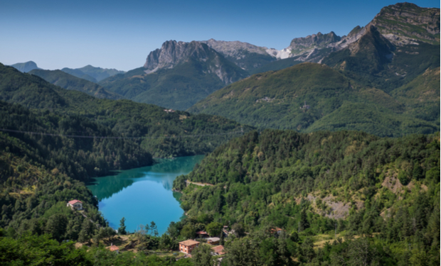 A turquoise lake sits nestled among lush green hills and dense forests, surrounded by rugged mountain peaks under a clear blue sky. Small houses are scattered in the valley below.