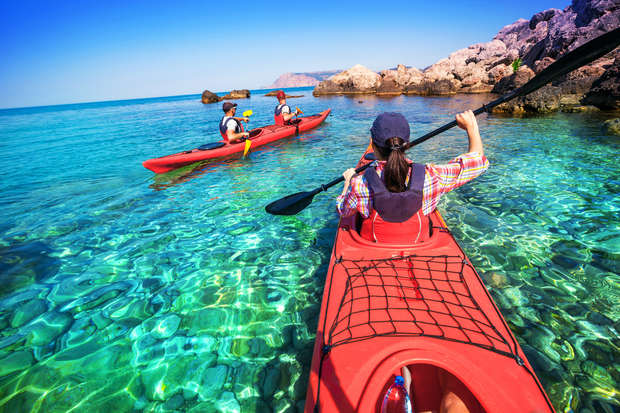 Kayakers paddle in clear, turquoise water near rocky shores under a bright blue sky. One kayaker is in the foreground, and two others are ahead in red kayaks.