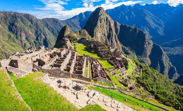 Ancient stone structures sit amidst lush green terraces, with towering mountain peaks in the background under a blue sky.