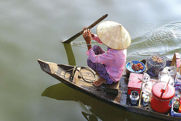 A person wearing a conical hat rows a wooden boat on a calm waterway, surrounded by containers of food and supplies, suggesting a floating market environment.