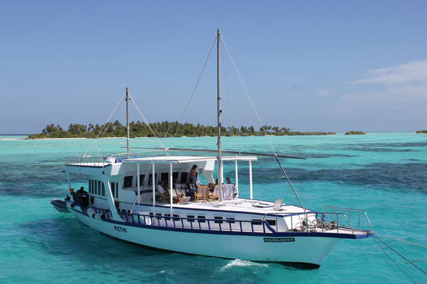 A white boat labeled "KEYIF" sails on clear turquoise water. Passengers relax on deck under a canopy with distant green islands and a clear blue sky in the background.