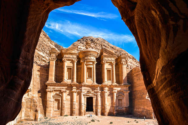 A grand, ancient stone facade with intricate carvings stands majestically under a bright blue sky, framed by dark rock walls resembling a cave entrance, in a desert landscape.
