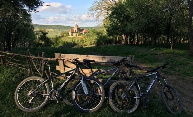 Three mountain bikes rest against a wooden fence in a grassy, wooded area. In the background, a church-like building with red roofs is visible under a blue sky with clouds.