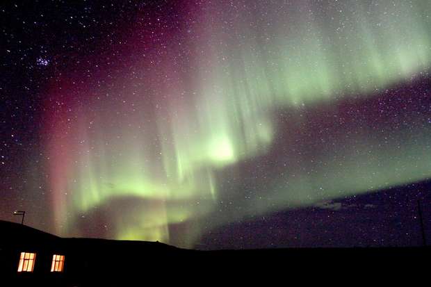 Aurora borealis displays colorful green and purple lights cascading in the night sky above a dark landscape. A house with illuminated windows is visible in the foreground.