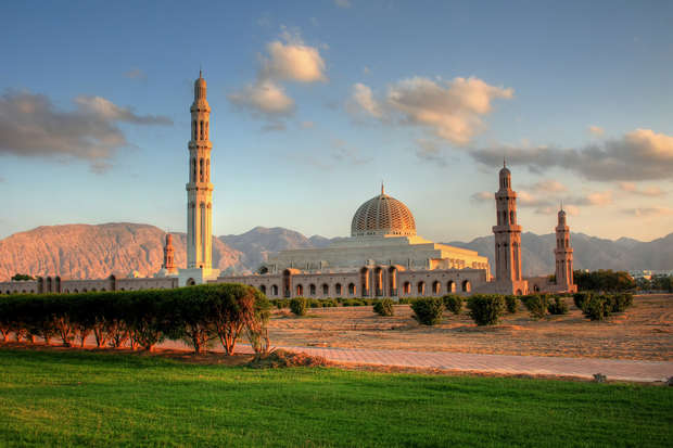A grand mosque with a large dome and tall minarets stands majestically, surrounded by a lush green field and mountainous backdrop under a partly cloudy sky at sunset.