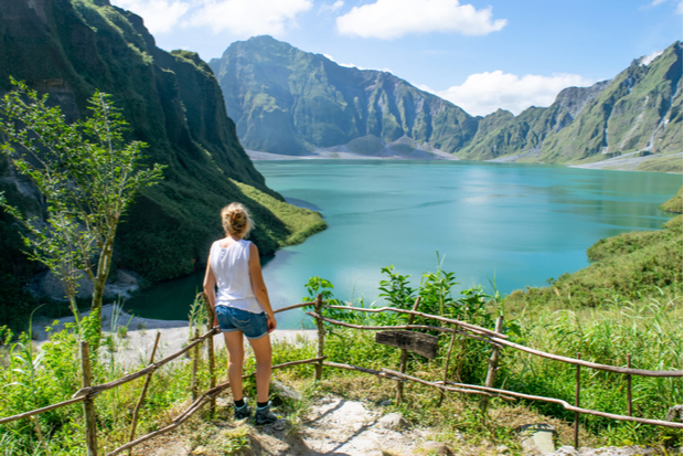 A person stands gazing at a tranquil, turquoise lake surrounded by towering, green mountains under a clear blue sky, with a rustic wooden fence and lush vegetation framing the foreground.
