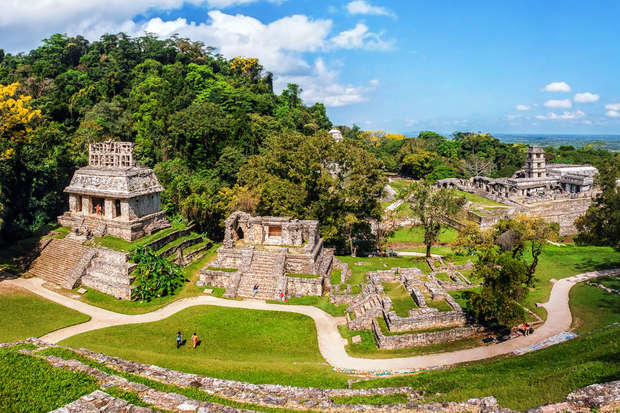 Ancient stone structures stand amid lush greenery, showcasing intricate architecture. Pathways weave between the ruins, with distant trees and a clear, blue sky framing the historical site.