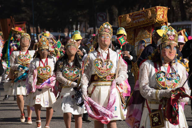 Dancers wear traditional, colorful costumes with detailed masks and embroidered vests, performing in a street parade amid a lively crowd.