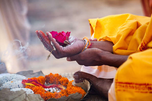 Hands hold a red rose and powdered substance, performing a ritual over a decorated surface with marigold flowers and smoke, surrounded by yellow fabric in a ceremonial setting.