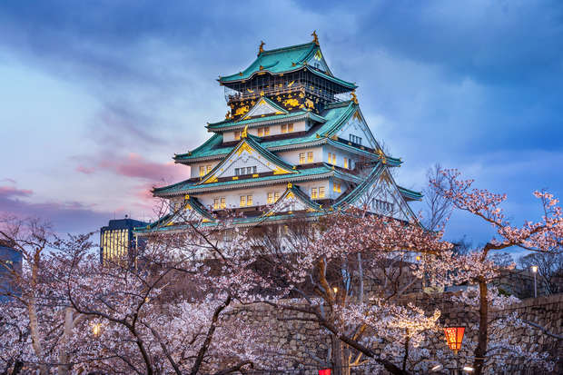 A large, traditional Japanese castle with ornate roofs sits amid blooming cherry blossom trees during dusk, under a dramatic, colorful sky.