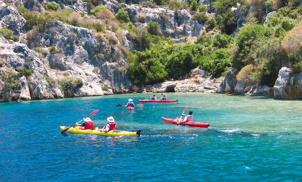 Kayakers paddle colorful boats through blue water near rocky cliffs with lush green vegetation, amid a sunny and clear coastal setting.