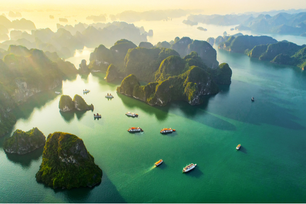 Boats float on emerald-green water surrounded by small, lush islands under a hazy golden sky, creating a serene, picturesque seascape.