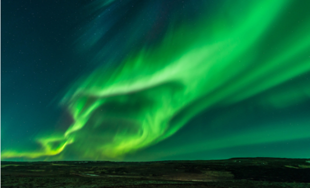 Vivid green auroras swirl dynamically across a dark night sky above a vast, open landscape, creating a striking natural spectacle with streaks of light and shadow.