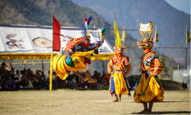 Dancers in colorful traditional costumes perform energetically, one leaping mid-air, with elaborate masks in an outdoor festival setting. Banners and a crowd are visible, set against a mountainous backdrop.