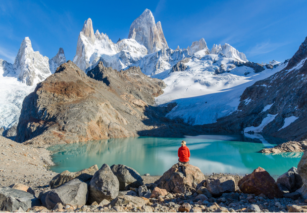 Person in a red jacket sitting on a rock, admiring a turquoise glacial lake with snow-capped mountains in the background under a clear blue sky.