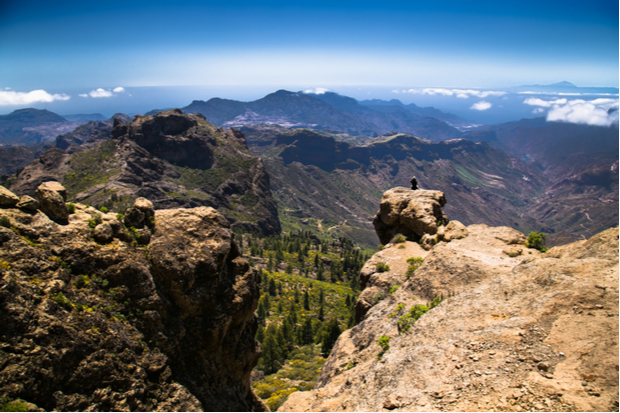A person sits on a rocky ledge, surrounded by vast mountain ranges and valleys under a clear blue sky. Sparse vegetation and some clouds enhance the rugged, natural landscape.