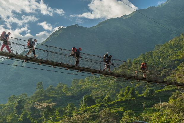 Hikers, with large backpacks, cross a suspension bridge above a lush, green valley surrounded by towering mountains under a cloudy sky.