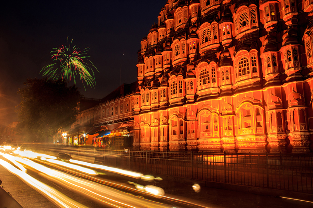 Ornate palace facade is illuminated at night, with colorful fireworks exploding above; streaks of light from passing vehicles blur on the street in the foreground.