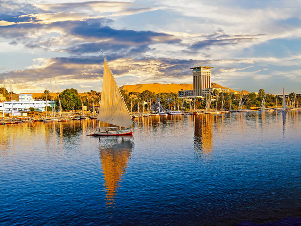 A sailboat glides across a reflective river under a dramatic sky. Tall buildings and lush greenery line the opposite shore, with a sandy hillside visible in the background.
