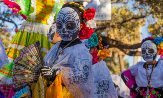 Individuals dressed in colorful traditional attire and ornate skull face paint hold fans, participating in a festive outdoor celebration amidst trees, capturing the vibrant spirit of Day of the Dead festivities.