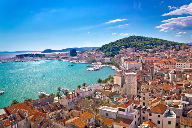 A coastal cityscape features turquoise water with boats; historic buildings with red roofs fill the foreground, and lush green hills and a blue sky provide a scenic backdrop.
