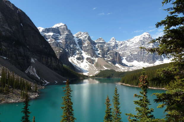 Turquoise lake reflects sunlight, surrounded by evergreen trees. Towering snow-capped mountains frame the scene under a clear blue sky, creating a serene and majestic natural landscape.