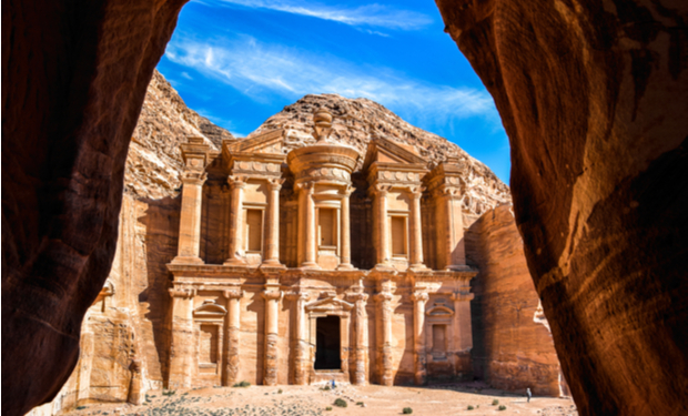 Ancient sandstone structure stands majestically, carved into a cliff, framed by natural rock formations; clear blue sky overhead enhances its grandeur in a desert setting.