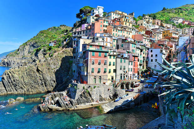 Colorful buildings perch on rocky cliffs, overlooking a crystal-clear blue sea, with boats anchored at the shoreline. Lush greenery covers the surrounding hills under a sunny, clear sky.