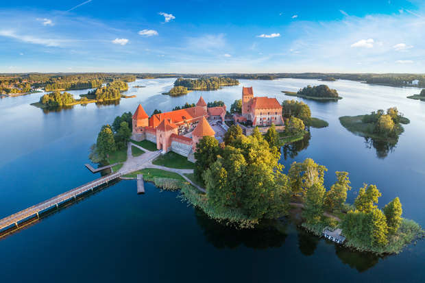 A historic castle with red roofs sits on an island, surrounded by serene blue water and lush greenery, connected to the mainland by a narrow wooden bridge under a clear sky.