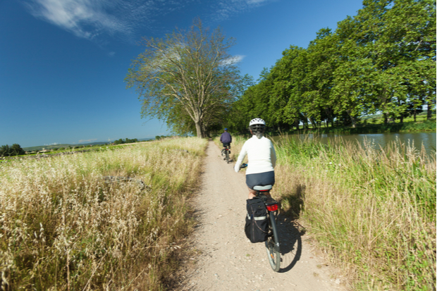 Two cyclists ride along a dirt path bordered by grassy fields and a tree-lined river under a clear blue sky.