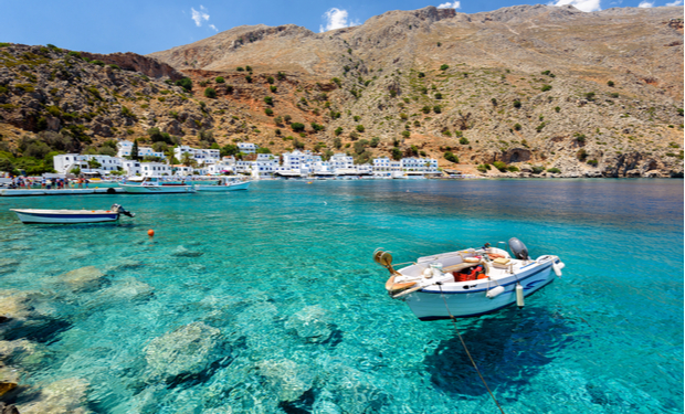 A small boat floats on crystal-clear turquoise water, docked near rocky shores. In the background, white houses line a rugged hillside beneath a bright blue sky.