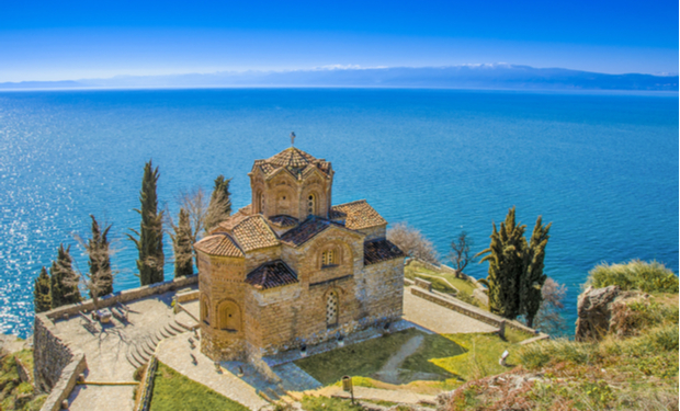 A historic stone church sits on a cliff, overlooking a vast, serene body of water with snow-capped mountains in the distance. Tall trees and a paved walkway surround the church.