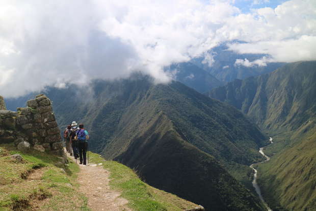 Hikers walk along a narrow mountain path, bordered by ancient stone walls, overlooking a lush green valley with distant peaks shrouded in clouds and a river winding through the landscape.
